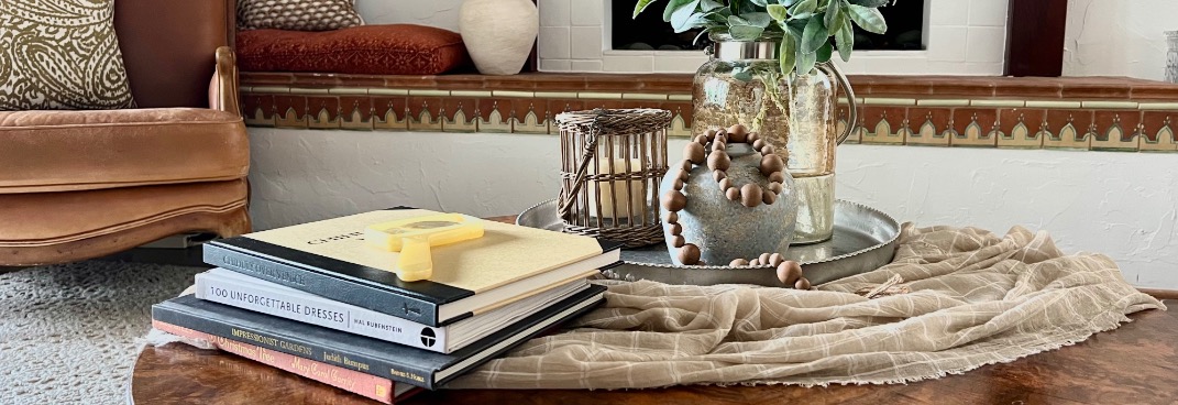 A cozy living room scene with a style round coffee table holding stacked books, a yellow toy camera, a decorative tray with a glass vase of greenery, wooden beads, and a wicker candle holder. A patterned sofa is in the background.