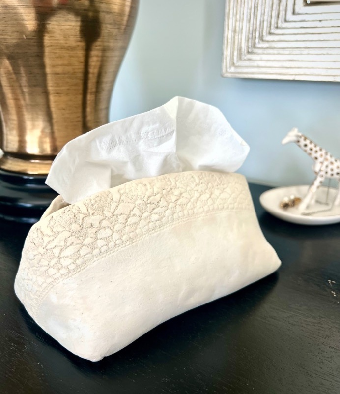 A white tissue box holder with lace detailing sits on a dark surface, next to a gold lamp and a small dish holding a decorative giraffe figurine.