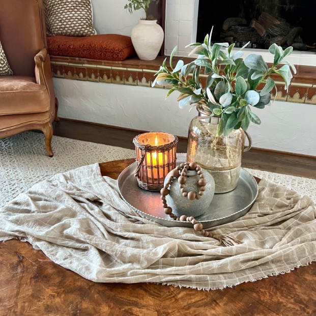 A cozy living room with a style round coffee table decorated with a light beige cloth, a candle in a glass holder, a grey vase with wooden beads, and a glass jar with green leafy branches on a metal tray.