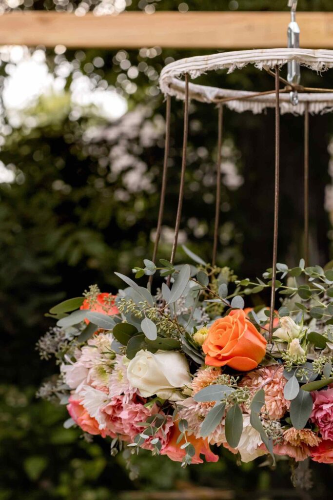 A floral chandelier with orange, white, and pink roses, greenery, and other flowers is hanging outdoors from a metal and rope frame. The background is blurred with trees and sunlight visible.