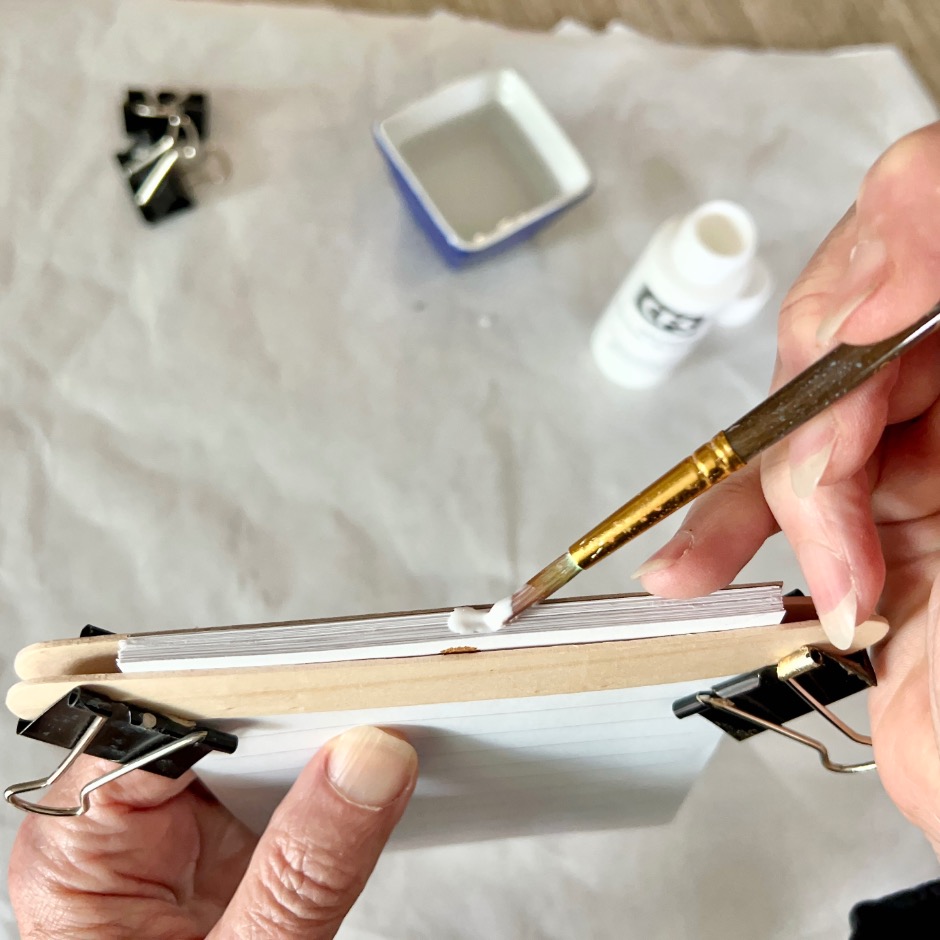 A hand uses a brush to apply white glue to the edge of a stack of notecards held together with binder clips and wooden sticks. A bottle of glue and a small dish are in the background on a sheet of white paper.