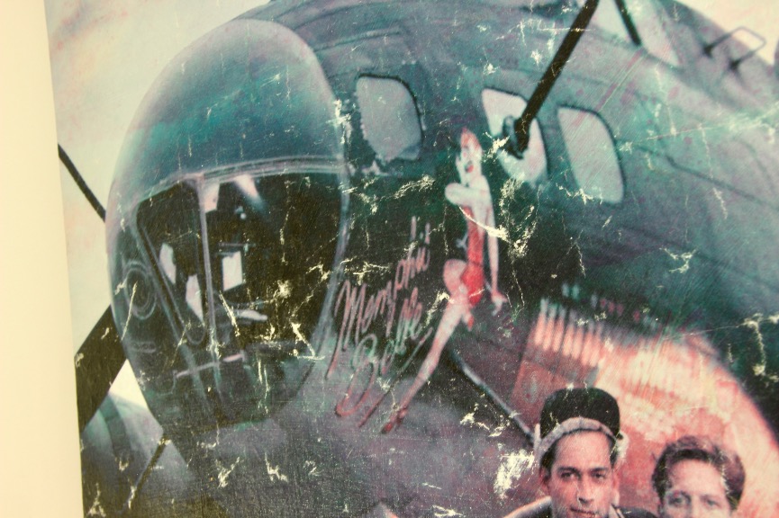 A close-up of a vintage military aircraft with worn paint, featuring nose art reminiscent of vintage poster artwork—the pin-up girl and “Myrtile Belle” near the cockpit—with part of a group visible in the lower right corner.