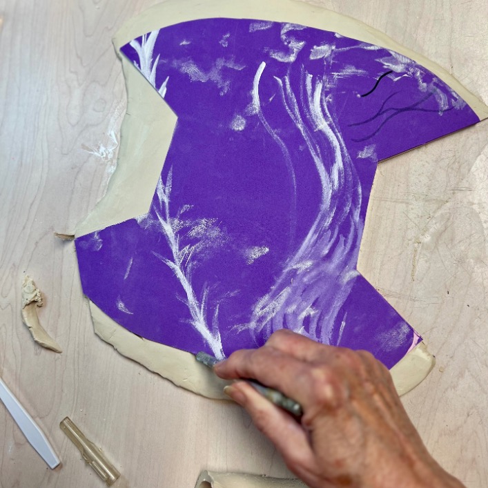 A hand uses a tool to draw white lines resembling tree branches on a purple surface attached to clay, crafting a diy tissue holder that rests on a light wooden table.