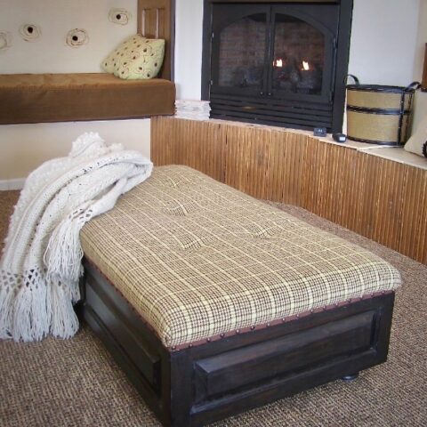 A cozy living room corner with a cushioned bench, a fireplace, a plaid upholstered ottoman, and a white knit blanket draped over it. The space features warm brown tones and a relaxed, inviting atmosphere.