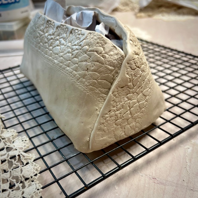 A handmade clay diy tissue holder with lace texture is drying on a cooling rack, paper stuffed inside to hold its shape. The surface beneath is covered with fabric and lace scraps.