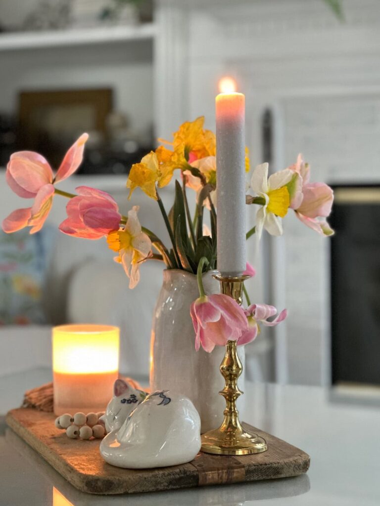 A lit white candle in a brass holder sits next to a ceramic vase with pink and yellow flowers, a glowing candle, and a small white bird ornament on a wooden tray atop a white table.