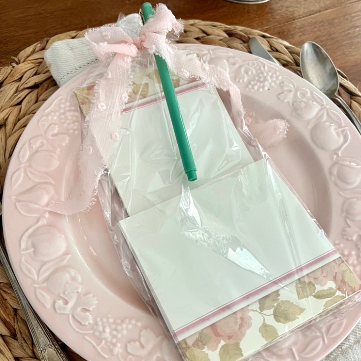 A floral notepad and green pen, wrapped in plastic and tied with a pink ribbon, sit on a pink decorative plate atop a woven placemat, surrounded by silverware.