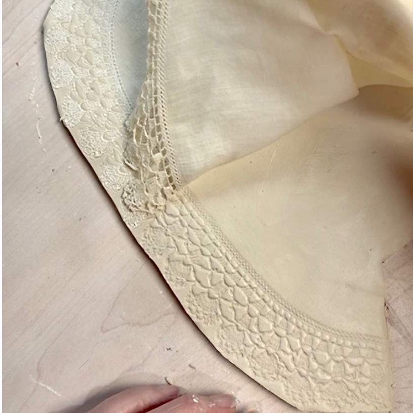 A close-up of a delicate, off-white fabric with intricate lace trim—perfect for a diy tissue holder. A hand is partially visible at the bottom, gently holding the cloth over a light-colored wood surface.