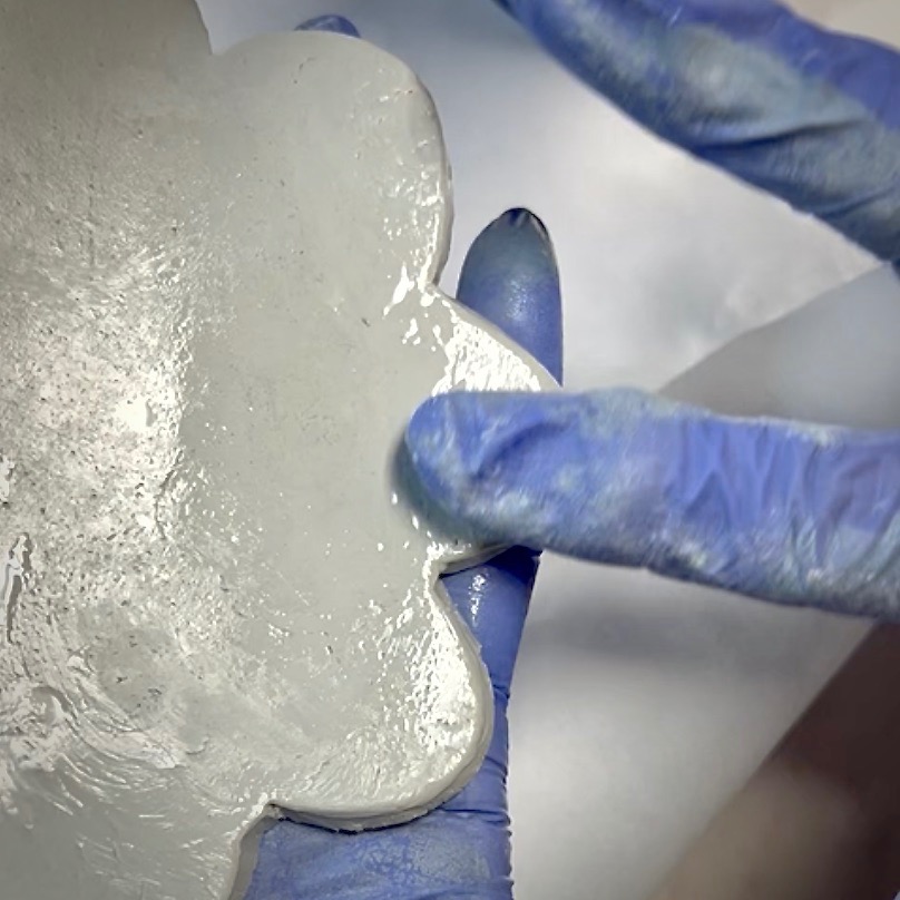 A close-up of two hands wearing blue gloves holding a wet, cloudy, flower-shaped piece of material, possibly clay or resin—perfect for a diy tissue holder—against a plain background.