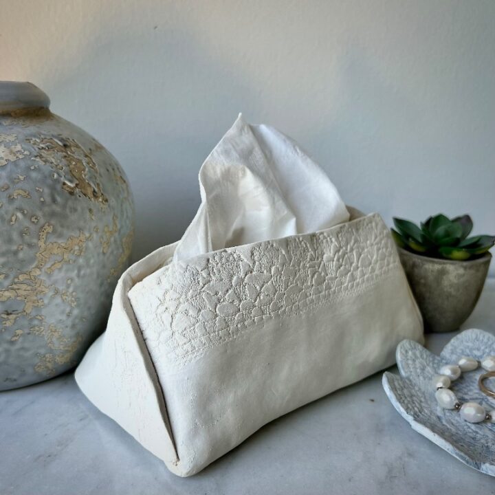 A white, textured DIY tissue holder sits between a decorative ceramic vase and a small potted succulent on a marble surface, with a beaded bracelet resting in a dish nearby.