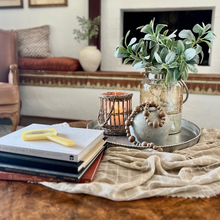 A cozy style round coffee table features a stack of books, a yellow magnifying glass, a folded cloth, and a metal tray holding a potted plant, beaded garland, and a lit candle in a wicker holder.