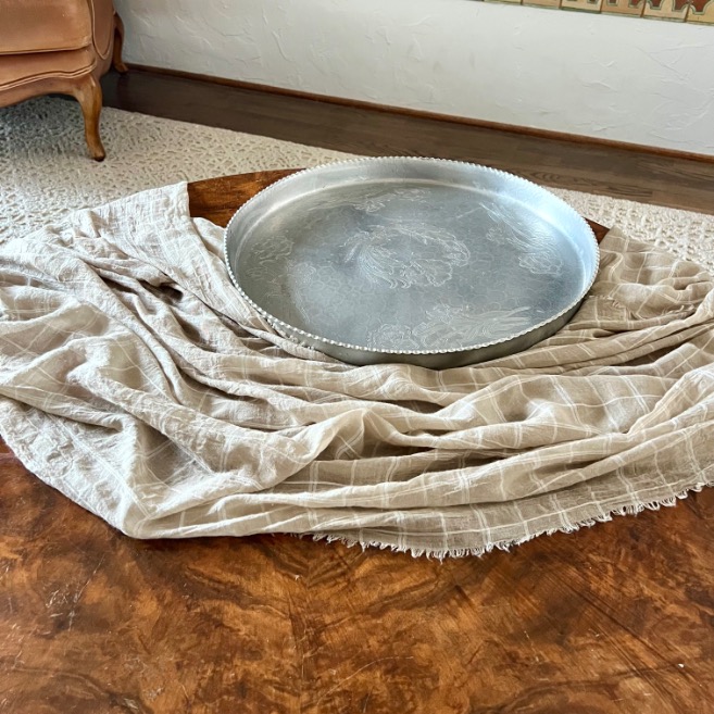 A large round silver tray rests on a beige checkered cloth draped across a wooden style round coffee table, with part of an upholstered chair and patterned rug visible in the background.