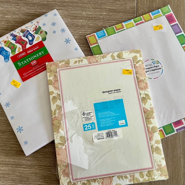 Three packs of decorative stationery paper with various borders—holiday stockings, pastel squares, and pink floral—are displayed on a wooden surface, each with a yellow price sticker.
