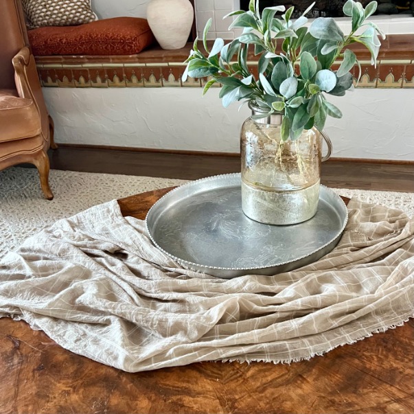 A large silver tray holding a clear glass jar with green leafy stems sits on a style round coffee table, draped with a beige cloth, in a cozy living room with neutral decor.