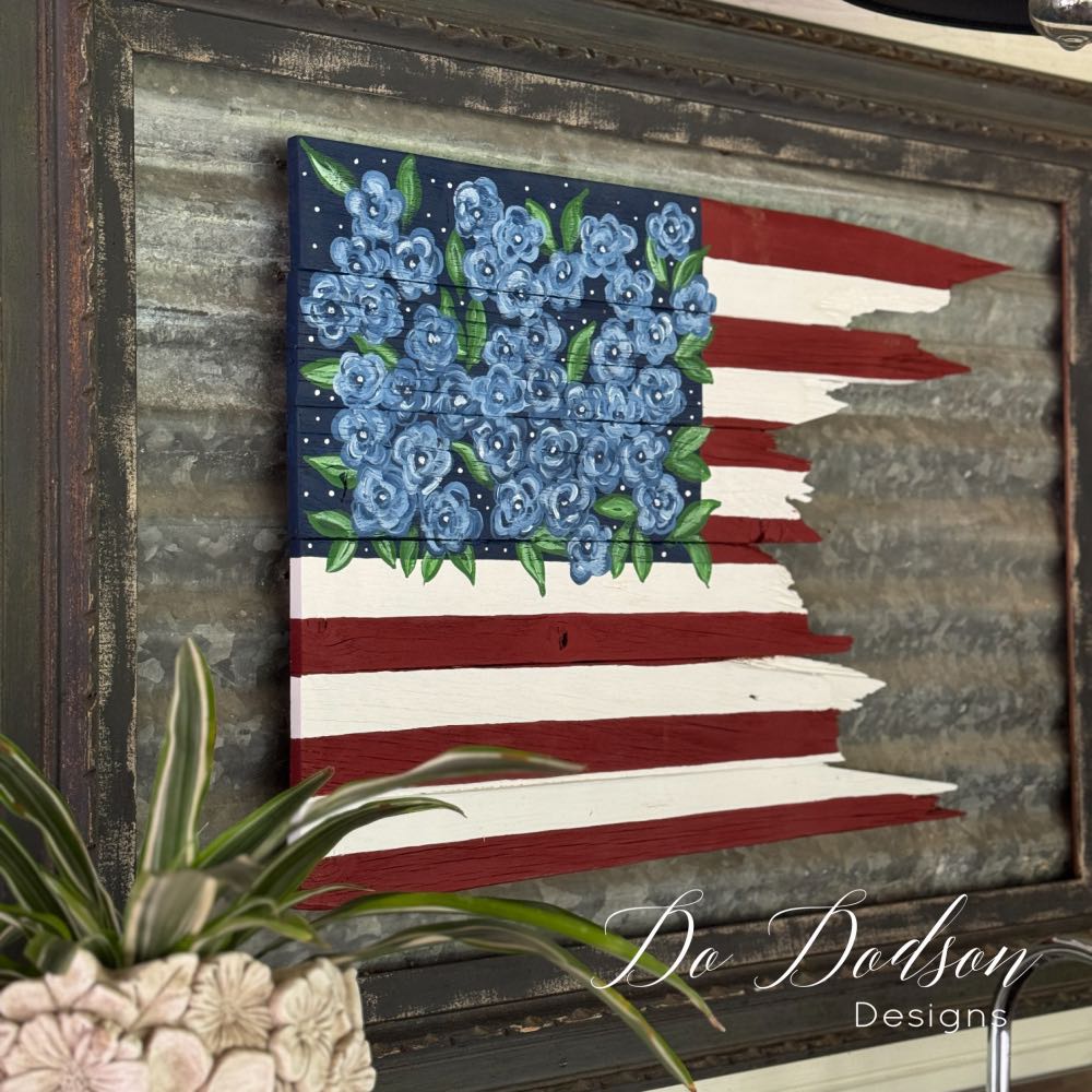 A rustic American flag art piece with red and white stripes and blue flowers in place of stars, mounted on wood. A green potted plant is in the foreground. "Do Dodson Designs" is written in the corner.
