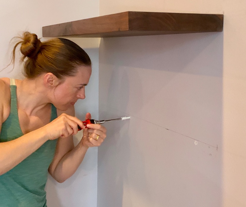 A woman in a green tank top uses a screwdriver to install a wall anchor beneath a wooden shelf, focusing intently as she prepares the space for floating bookshelves.
