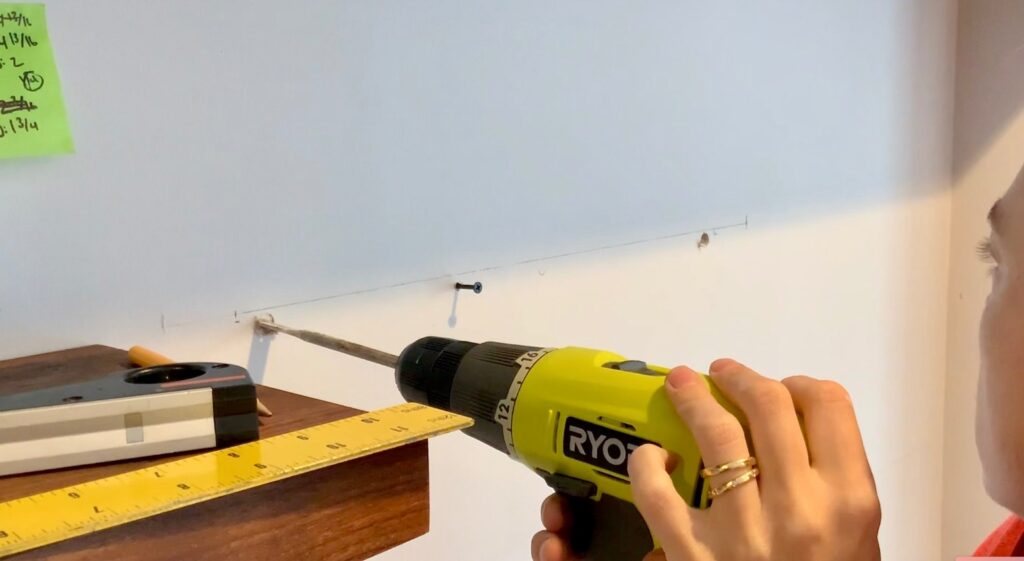 A person uses a yellow Ryobi power drill to screw into a wall above a wooden shelf, likely preparing to install floating bookshelves. A ruler, level tool, and sticky notes with writing are visible nearby.