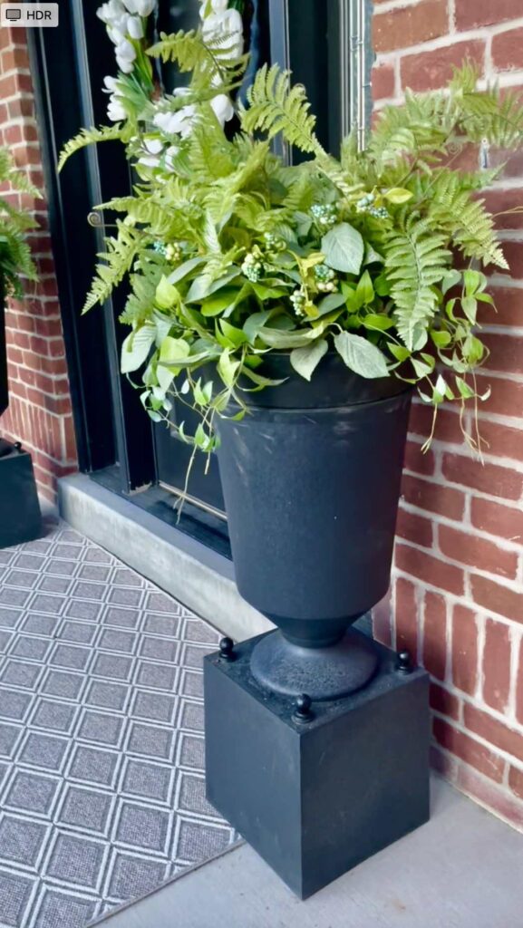 A black planter with green ferns and leafy plants sits on a black pedestal by a brick wall and glass door, next to a gray patterned outdoor rug.