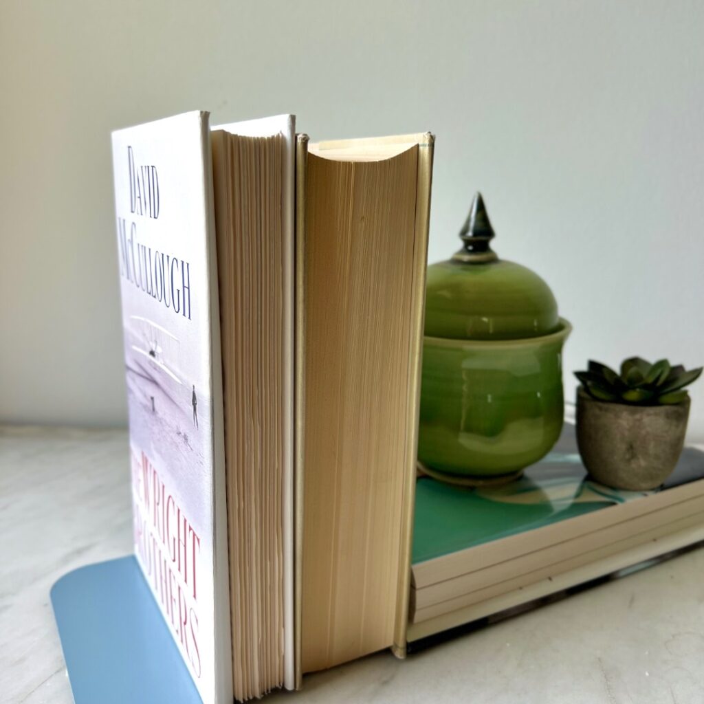 Two upright books between blue metal bookends, with a green ceramic jar and a small potted succulent resting on a horizontal book—all arranged as if on floating bookshelves atop a white marble surface.