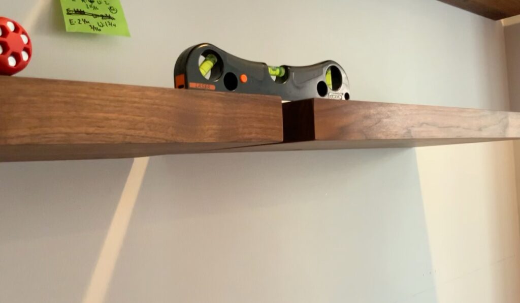 A black and green level tool sits on two misaligned floating bookshelves mounted on a wall; one shelf is visibly higher than the other. A red object and a green sticky note are partially visible on the left.