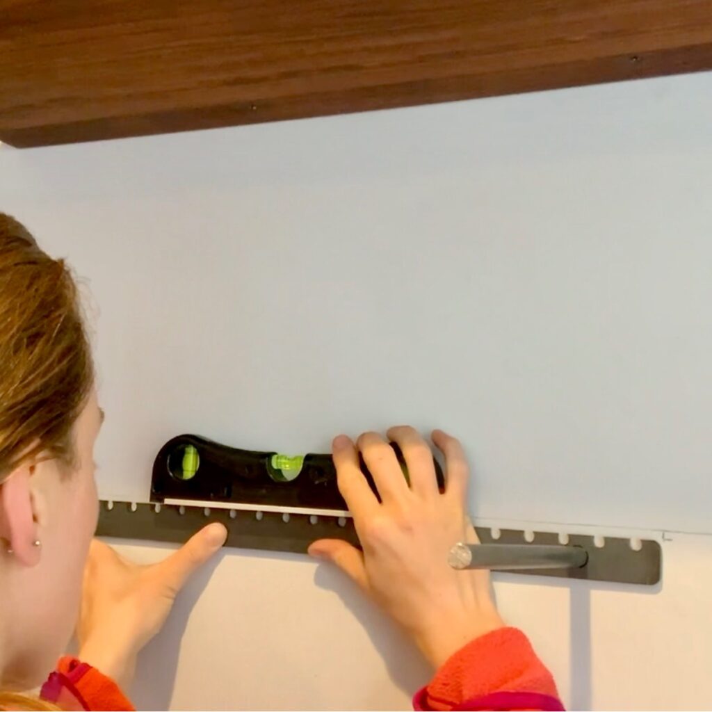 A person uses a spirit level to check the alignment of a metal bracket on a white wall, preparing to mount floating bookshelves under a wooden shelf.