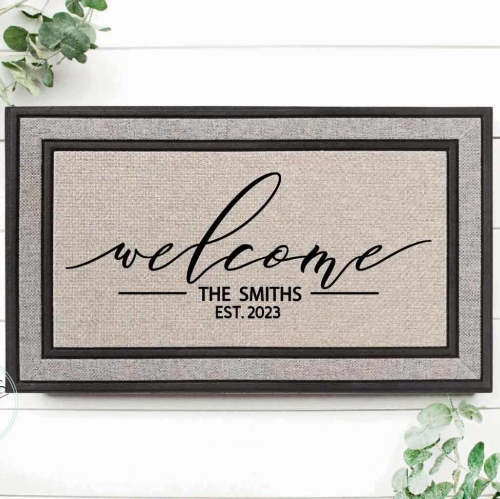 A doormat with black text that reads “welcome THE SMITHS EST. 2023” on a beige background, framed by a black border, surrounded by green leaves on a white wooden surface.