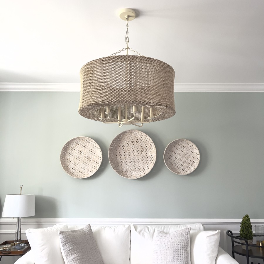 A living room with a woven chandelier, three decorative round wall plates, a white sofa with two pillows, a table lamp, and a small topiary on a side table against a light gray wall.
