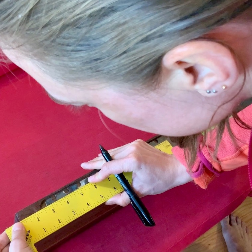 A person with light brown hair is measuring and marking a piece of wood with a yellow ruler and black pen on a red surface, preparing materials for floating bookshelves. Their hand holds the ruler steady while they draw a line.