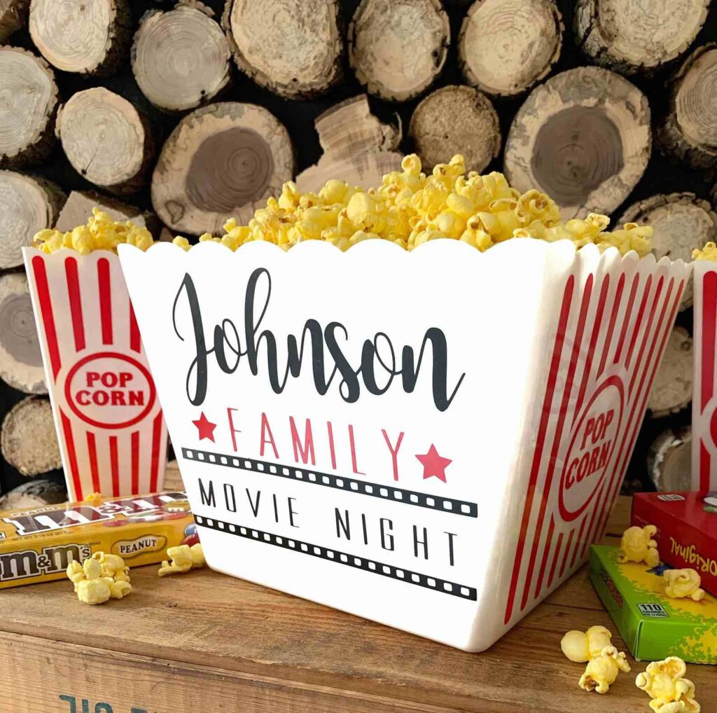 A large popcorn container with "Johnson Family Movie Night" printed on it, filled with popcorn, sits on a wooden surface. Candy boxes and popcorn kernels are nearby, with stacked firewood in the background.