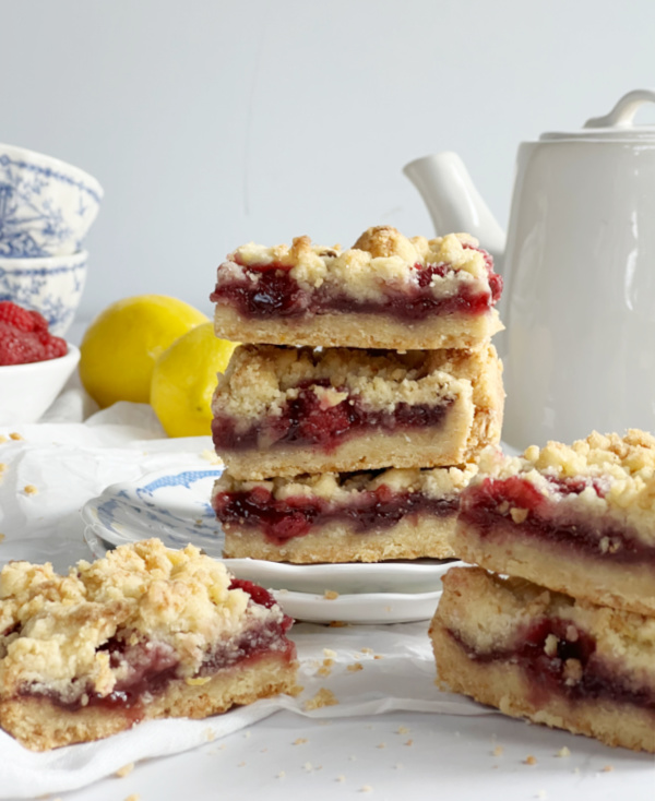 Three stacked crumb bars filled with strawberry and lemon, surrounded by more bars. In the background, lemons, a teapot, blue-and-white dishes, and a bowl of strawberries are visible.
