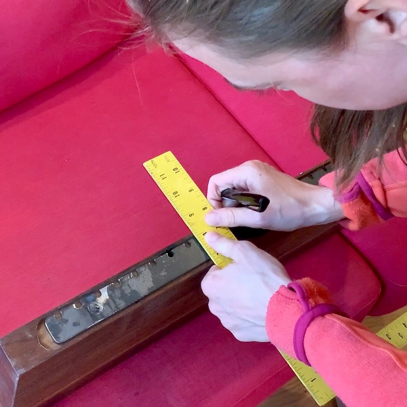 A person in a pink sweater uses a yellow ruler and a black marker to measure and mark a wooden part of a red upholstered couch, preparing pieces for building floating bookshelves.
