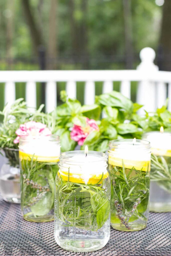 Three mason jars filled with water, lemon slices, fresh herbs, and floating candles sit on an outdoor table, with green plants and pink flowers blurred in the background.