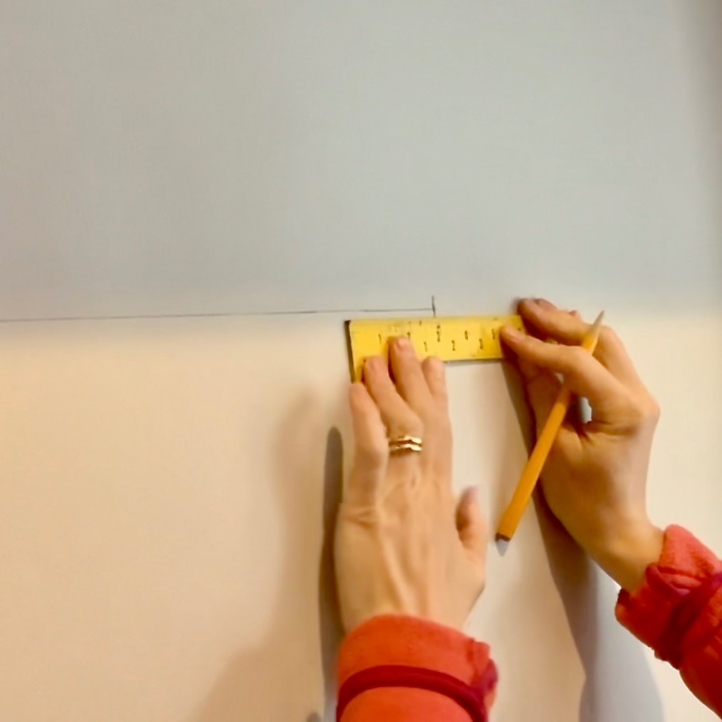 Two hands use a yellow measuring tape and pencil to mark a point on a light-colored wall, preparing to install floating bookshelves. The person is wearing a red long-sleeve shirt and a gold ring.