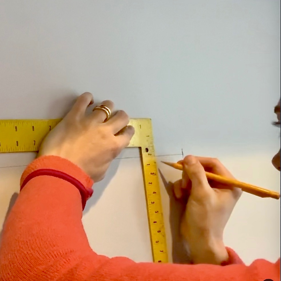 A person wearing a red sleeve uses a yellow ruler and pencil to measure and mark a straight line on a white wall, preparing to install floating bookshelves. Only their hands and part of their face are visible.