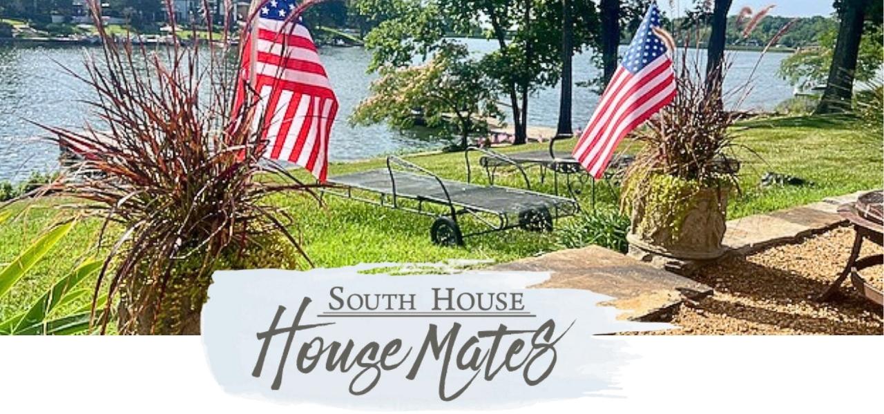 Two American flags flank a metal bench overlooking a lakeside lawn with trees and water in the background. Text below says "South House House Mates" in decorative lettering.