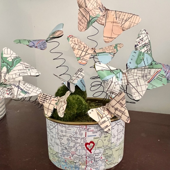 A creative idea for what to do with old maps: colorful map paper butterflies flutter on black wire spirals in a map-covered container with a red heart, all nestled in artificial moss.