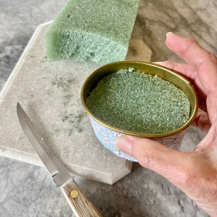 A hand holds an open tin can filled with green floral foam, as a knife and a block of the same foam rest on a marble cutting board—an upcycling idea as creative as exploring what to do with old maps.