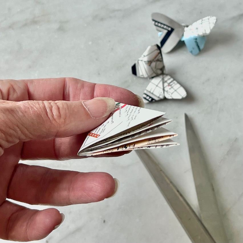A hand holds folded paper, preparing to cut; scissors and two finished paper butterflies made from map pages suggest a creative idea for what to do with old maps, all displayed on a white surface in the background.