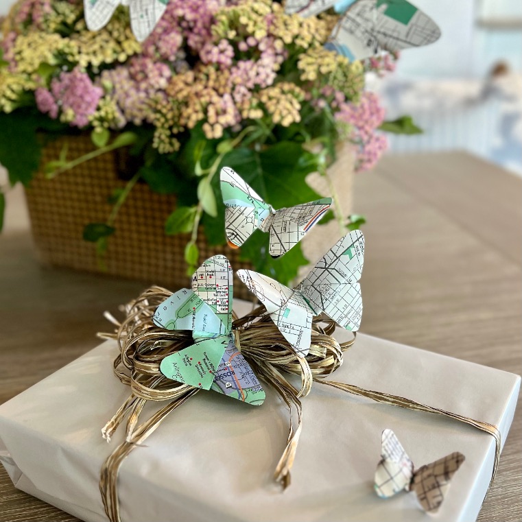 A white gift box is decorated with map-patterned paper butterflies, offering a charming idea for what to do with old maps. In the background, a woven basket holds pink and yellow flowers on a wooden surface.