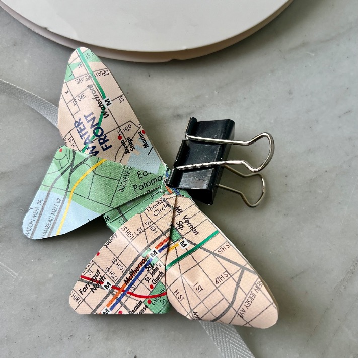 A butterfly-shaped paper craft made from a city map page—perfect if you’re wondering what to do with old maps—is held together with a black binder clip, resting on a gray surface.