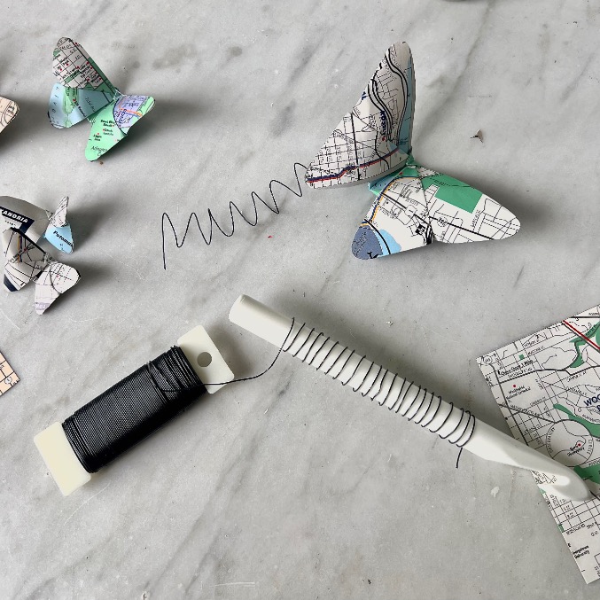 Give old maps new life: paper butterflies made from maps, a spool of black thread, a white craft tool, and coiled wire are artfully arranged on a white marble surface—perfect inspiration for what to do with old maps.