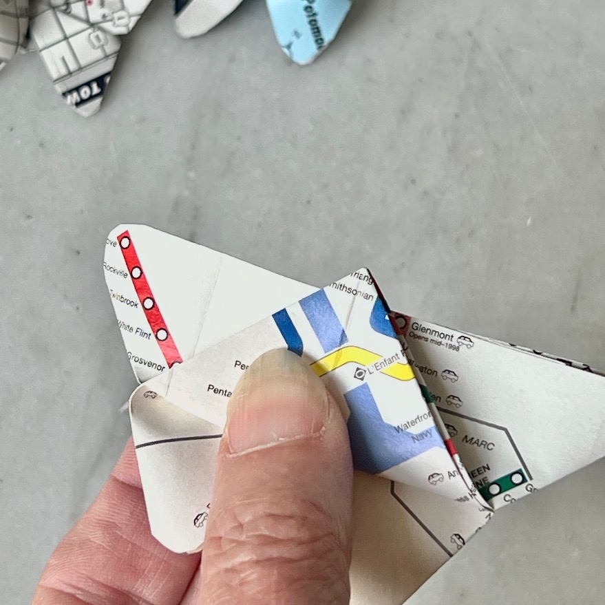A hand holds a paper butterfly folded from a map, suggesting what to do with old maps. Other similarly folded butterflies rest on a light gray surface in the background.