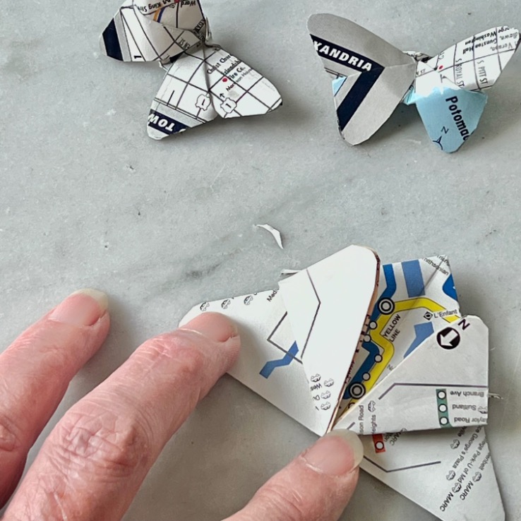 What to do with old maps: Fold them into beautiful butterflies, as shown by a hand crafting a butterfly from map paper, with two finished map butterflies resting on a light gray surface nearby.