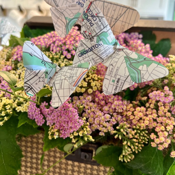 Paper butterflies made from old city maps are perched on a bouquet of pink and yellow flowers, with leafy greenery and a blurred background—an inspiring idea for what to do with old maps.