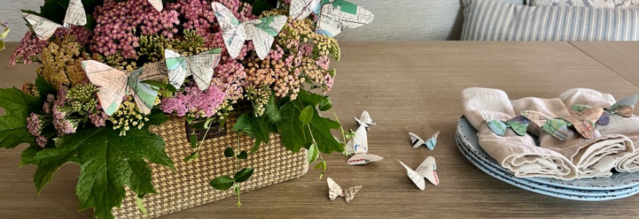 A woven basket filled with pink and yellow flowers, decorated with paper butterflies made from old maps. On a wooden table nearby are stacked plates, napkins, and more map-page butterflies on napkin rings—great ideas for what to do with old maps!.