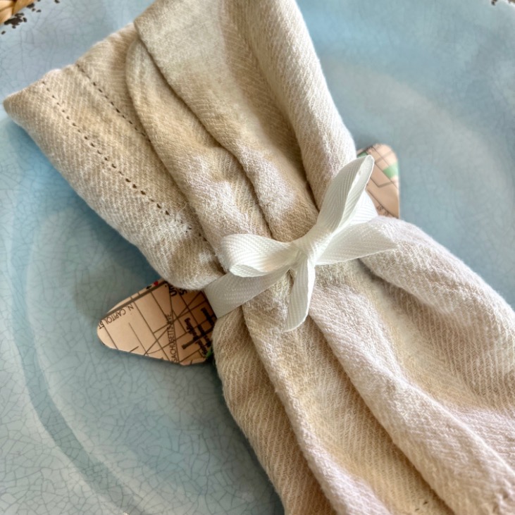 A cloth napkin wrapped with a white ribbon, tucked with a small paper heart made from old maps—ideal inspiration for what to do with old maps—placed on a light blue textured plate.