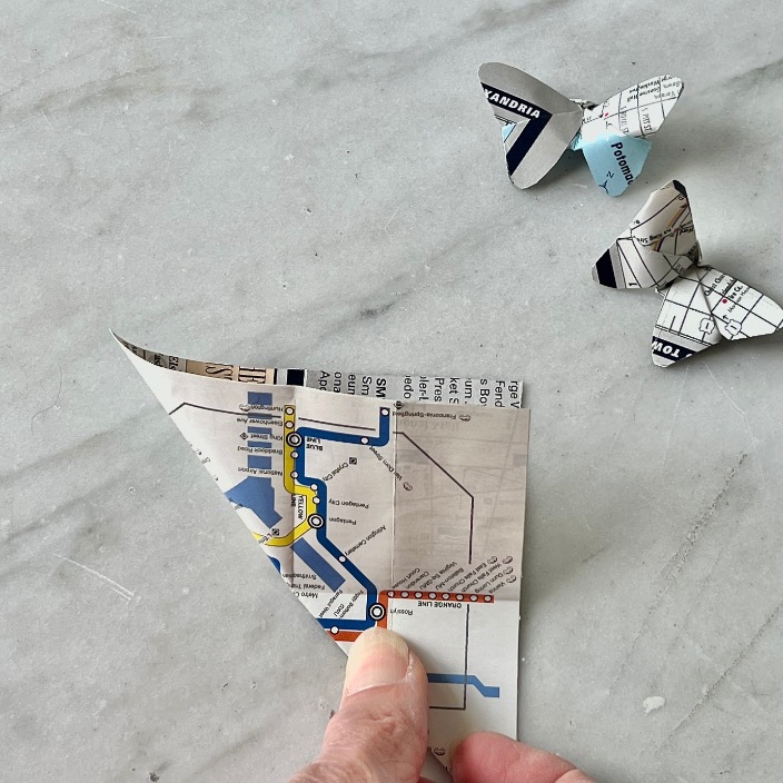 A hand folds a piece of a subway map into an origami butterfly on a grey surface, with two completed paper butterflies in the background—an inspiring idea for what to do with old maps.