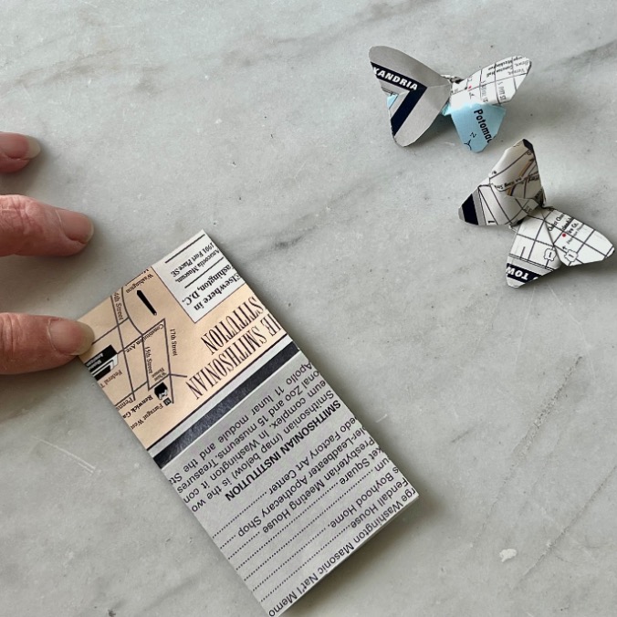 A hand touches a folded piece of paper with printed text and maps. Nearby, two small paper butterflies, crafted from old maps, rest on a light-colored surface—showing what to do with old maps in creative ways.
