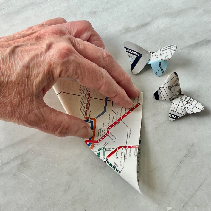 A hand is folding a piece of map paper, showcasing what to do with old maps, while two butterflies crafted from similar map paper rest on a marble surface nearby.