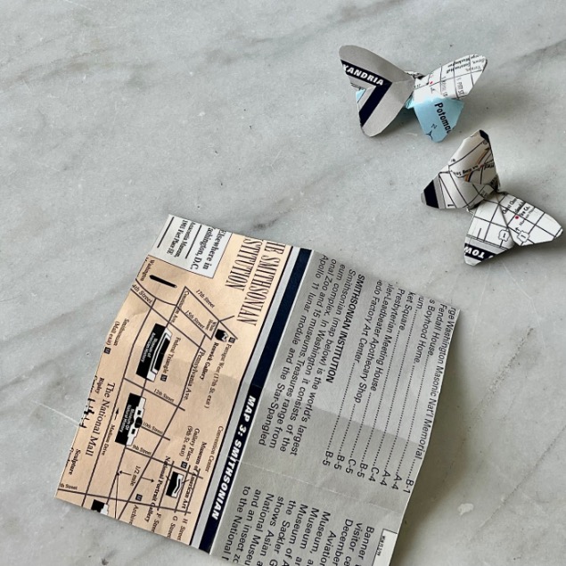 A piece of vintage map paper and two small butterflies cut from map paper are placed on a light gray marble surface, showing a creative idea for what to do with old maps—the butterflies appear to be flying away from the map piece.